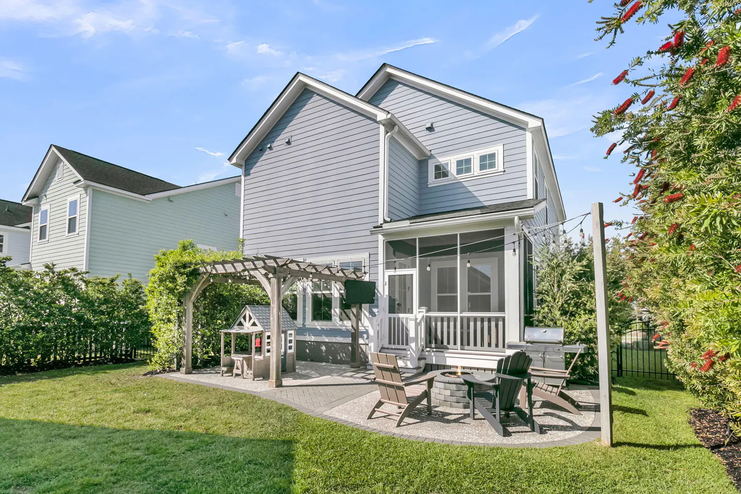Backyard of a gray house with a patio, fire pit, chairs, grill, and a playhouse under a pergola.