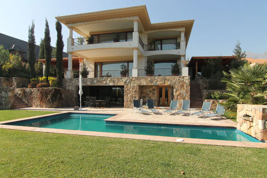 Two-story house with a pool. The house is white with stone accents and a balcony. Lounge chairs sit by the turquoise pool on green grass.