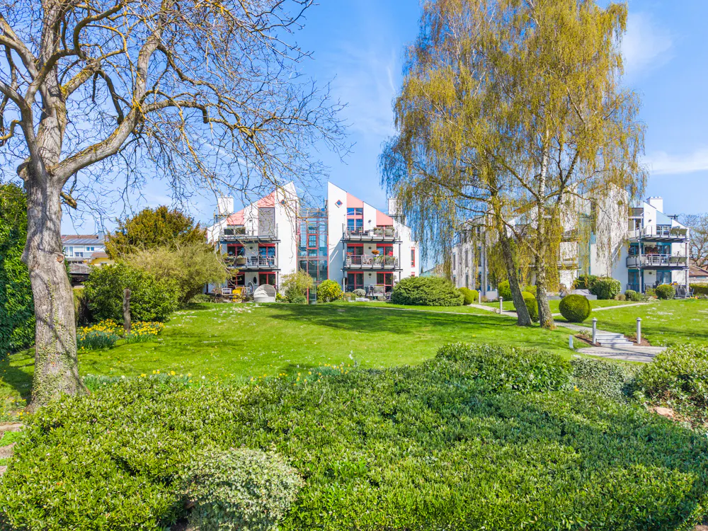 Exterior view of modern white apartments with balconies, surrounded by green lawns and trees under a blue sky.