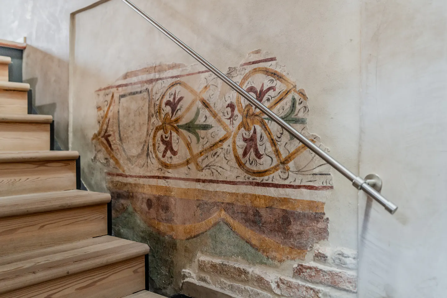 Wooden staircase next to a wall featuring exposed brick and a faded, decorative historical fresco with a metal handrail.