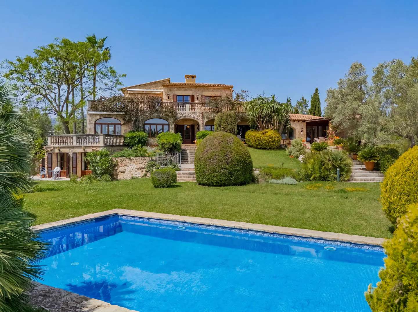A large stone house with a blue tiled pool in the foreground on a sunny day.