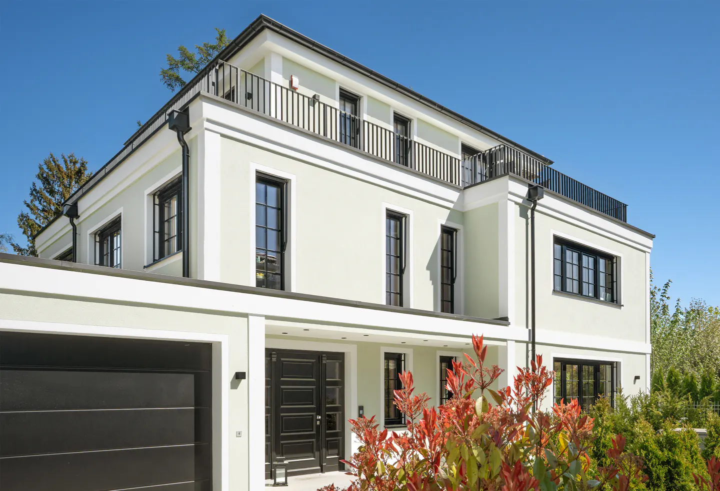 Exterior view of a three-story, light green house with black windows, a black garage door, and a black balcony railing.