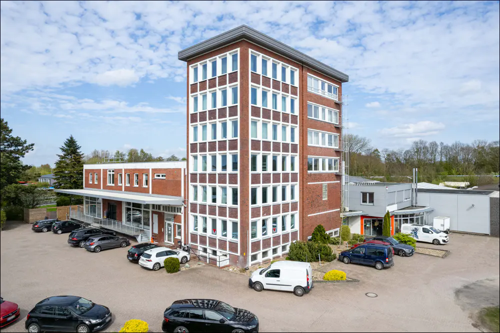 A brick building with white-framed windows and a parking lot filled with cars on a sunny day.