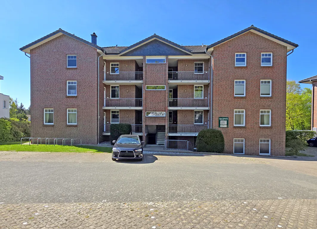 Three-story red brick apartment building with balconies, a central entrance, and a parked car in the paved front lot.
