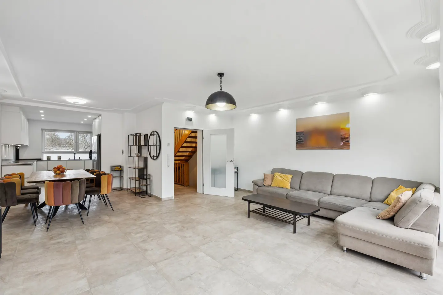 Open-concept living space with a gray sectional sofa, dining table with colorful chairs, and a staircase in the background.