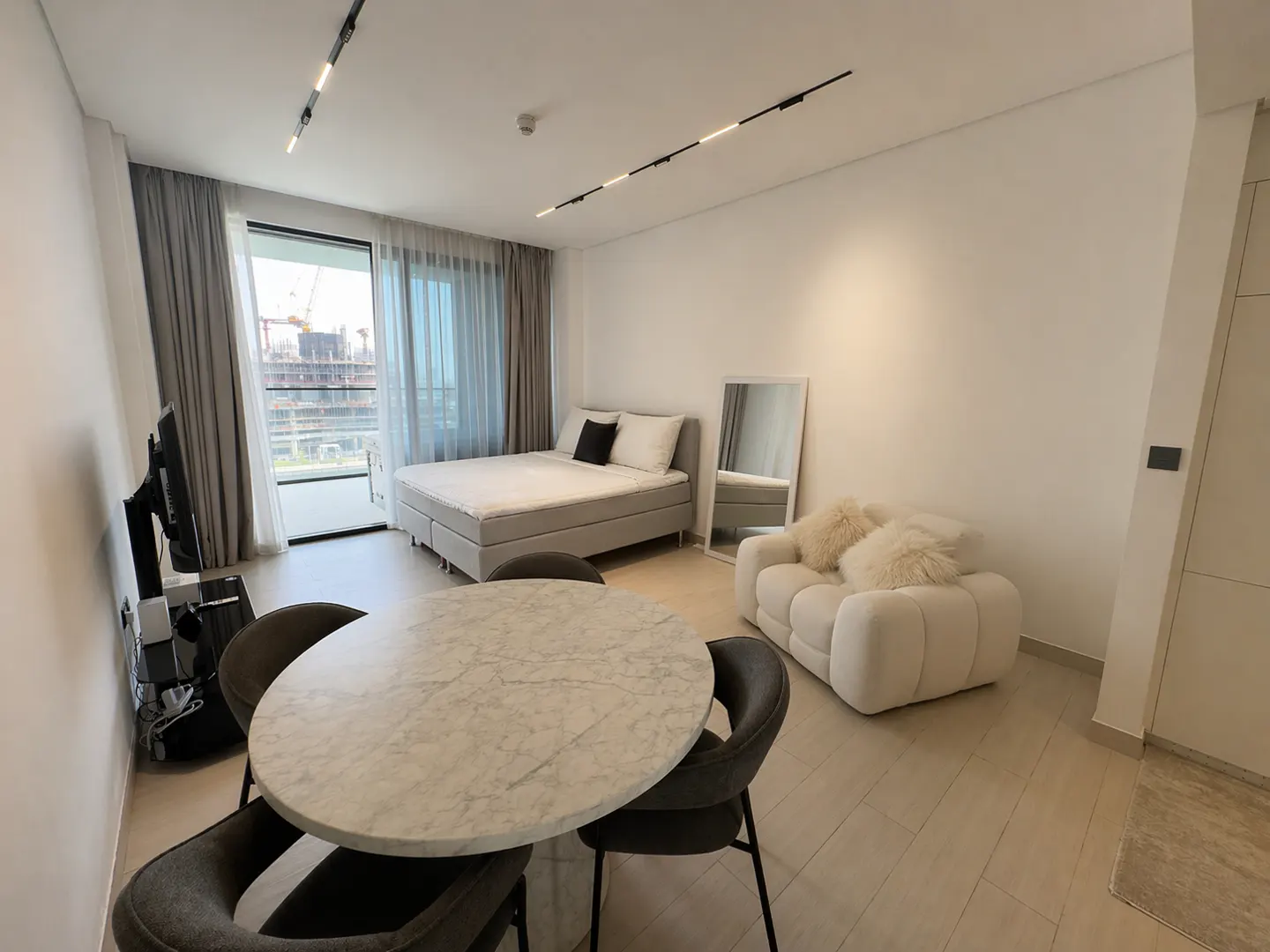 A bright studio apartment with a bed, sofa, table, and chairs. A large window overlooks a construction site.