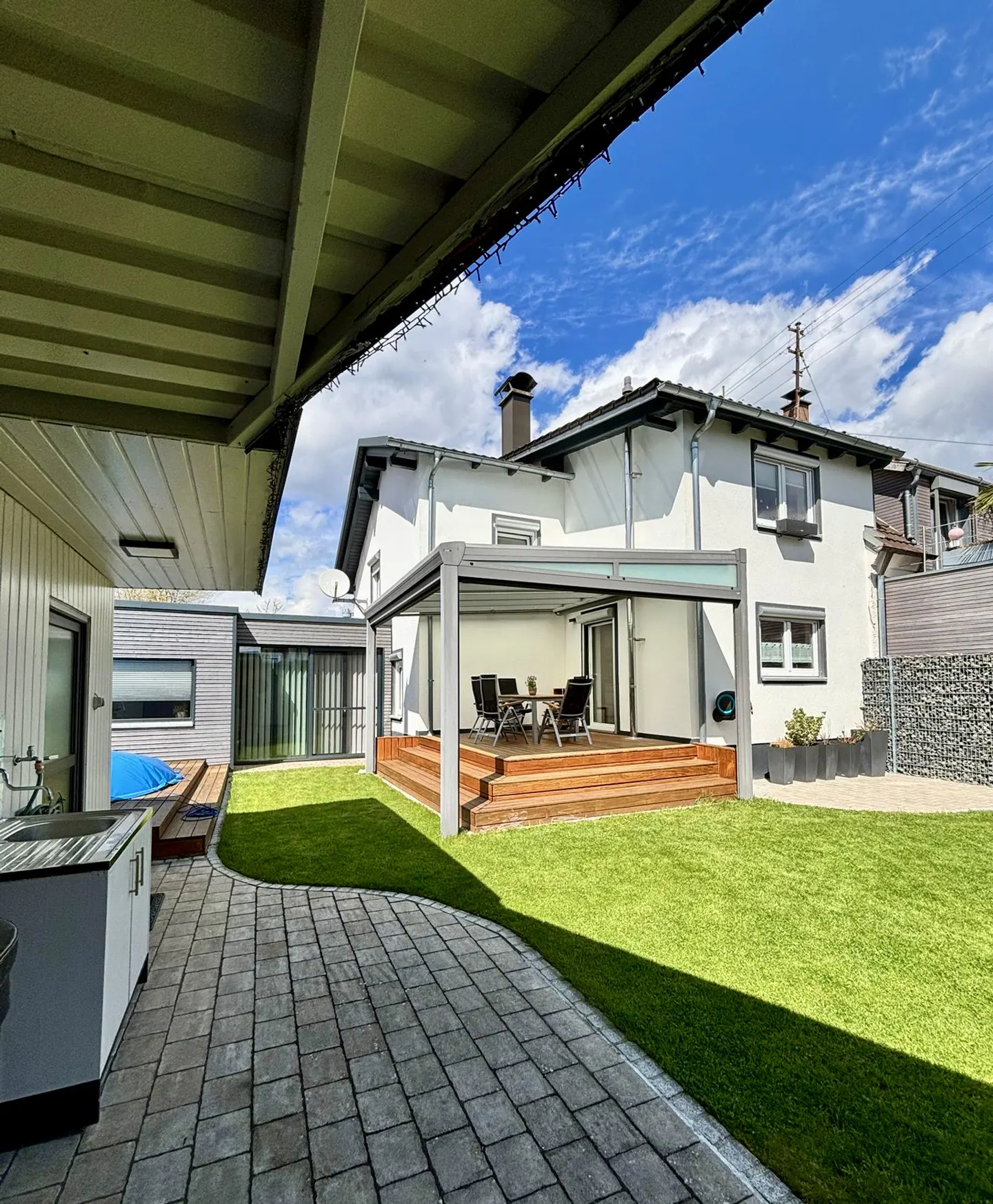 A backyard with a patio, green grass, and a white two-story house under a blue sky.