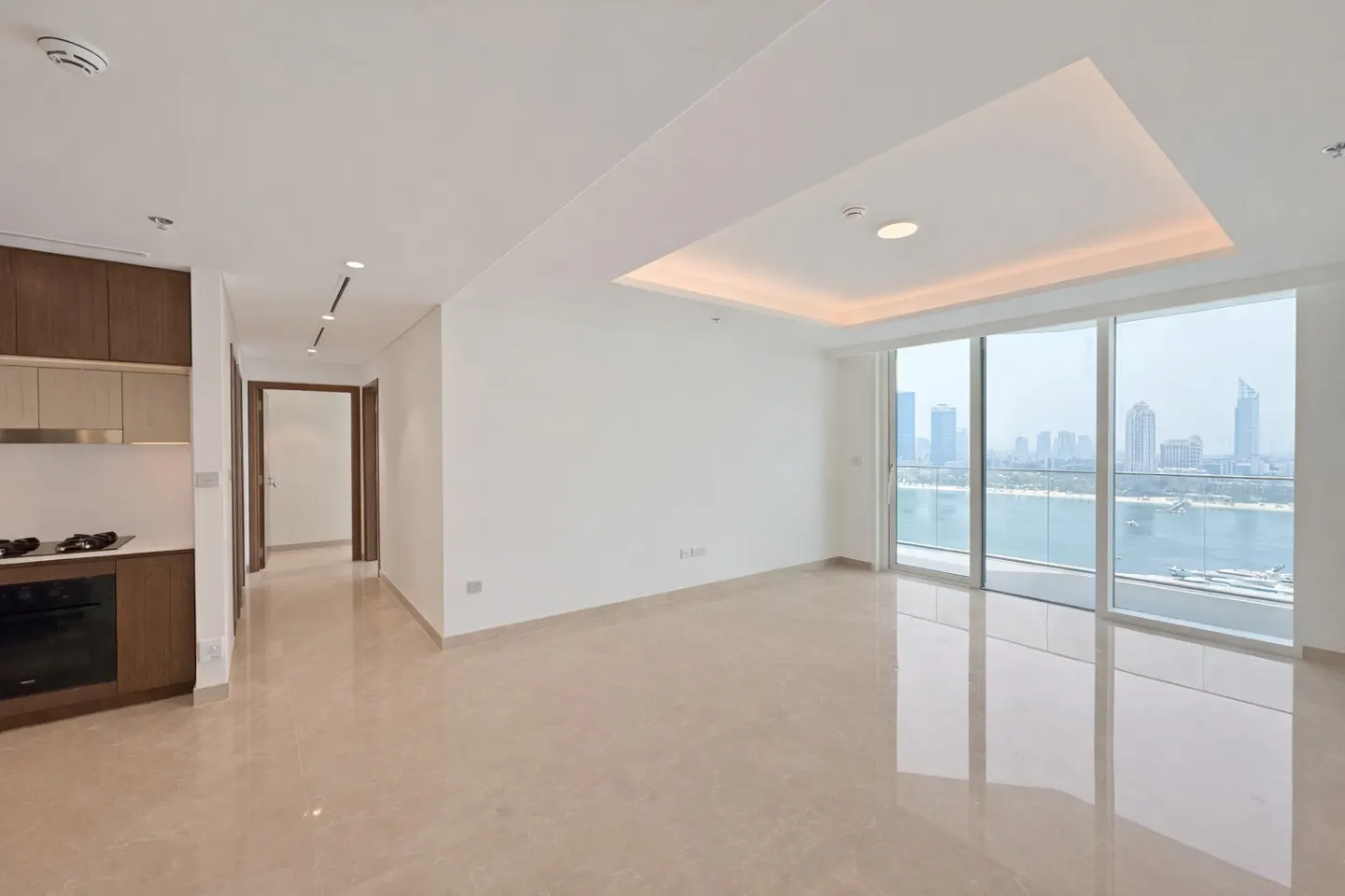 Bright, empty apartment with beige tile floors, white walls, and a kitchen area. Large windows offer a city and ocean view.