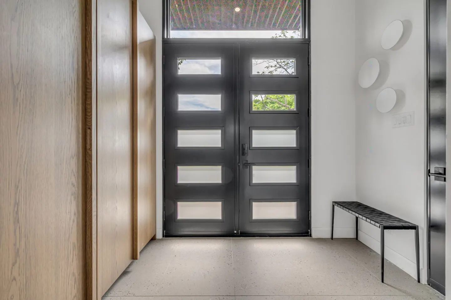 Modern entryway with a black double door, light wood cabinets, a black bench, and white walls with circular wall art.