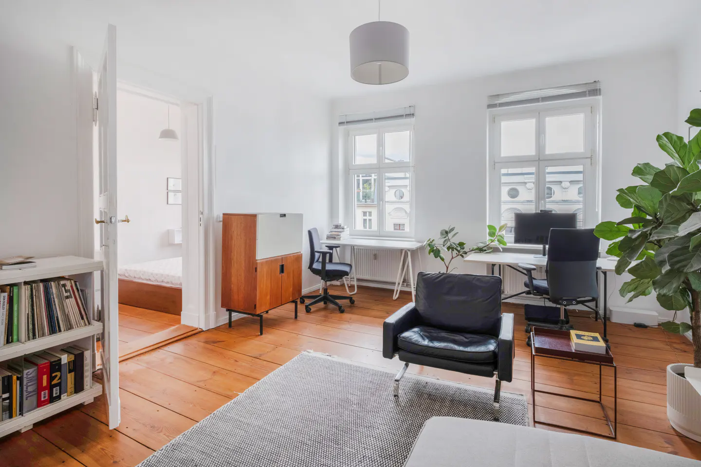 Bright, white home office with wood floors, two desks, black chairs, and a black leather armchair. A bookshelf and bedroom are visible through an open doorway.