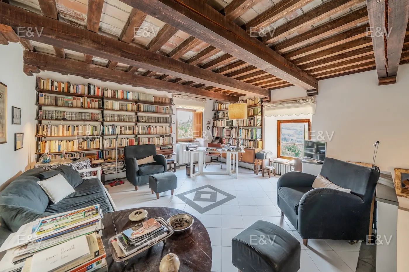 A cozy living room with floor-to-ceiling bookshelves, dark armchairs, and a wooden-beamed ceiling.