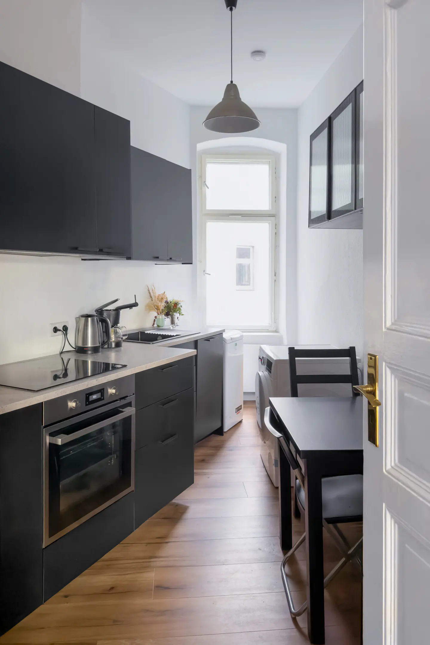 Modern kitchen with black cabinets, wood floors, and white walls. A table and chairs sit near a window with laundry machines.
