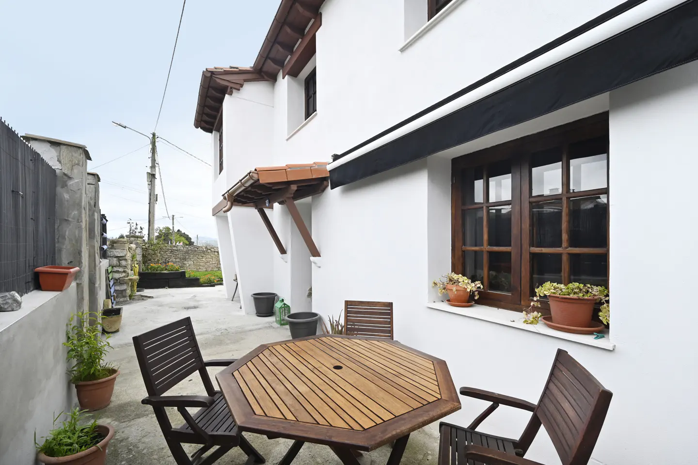 Outdoor patio with wooden table and chairs, potted plants, and a white building with brown window frames and awning.