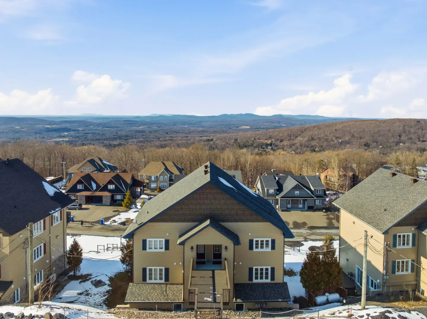 Aerial view of a tan two-story house with a brown roof, surrounded by other houses and a wooded landscape under a blue sky.