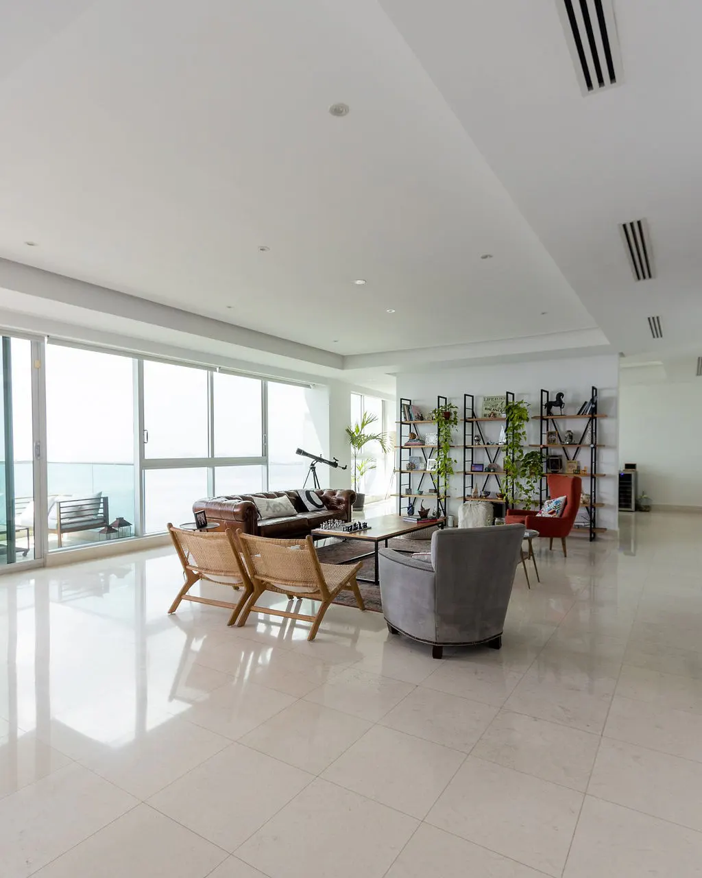 Bright, airy living room with ocean view. Brown leather sofa, woven chairs, and bookshelves with plants. White tile floor reflects light.