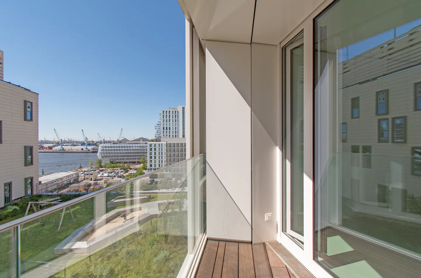 Balcony view of a city harbor. Glass railing, wood floor, white walls. Cranes and buildings in the background under a blue sky.