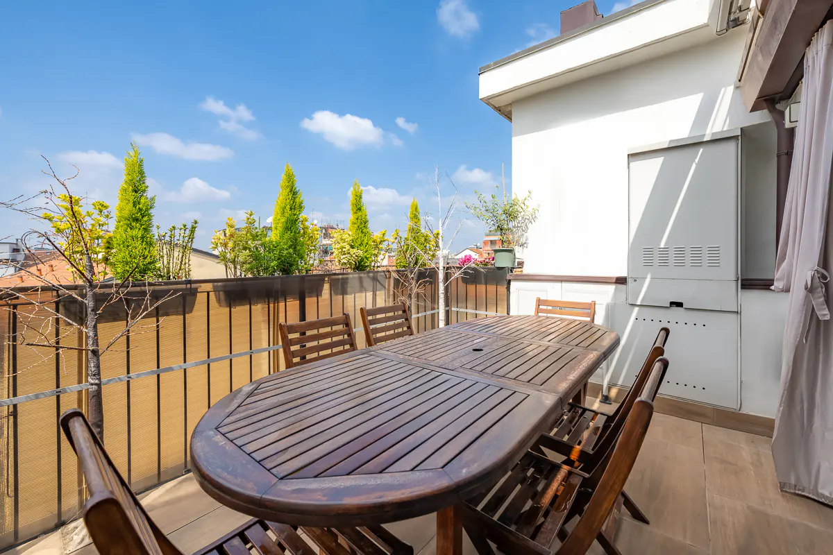 Outdoor patio with a wooden table and chairs, a metal railing, and green plants against a blue sky.