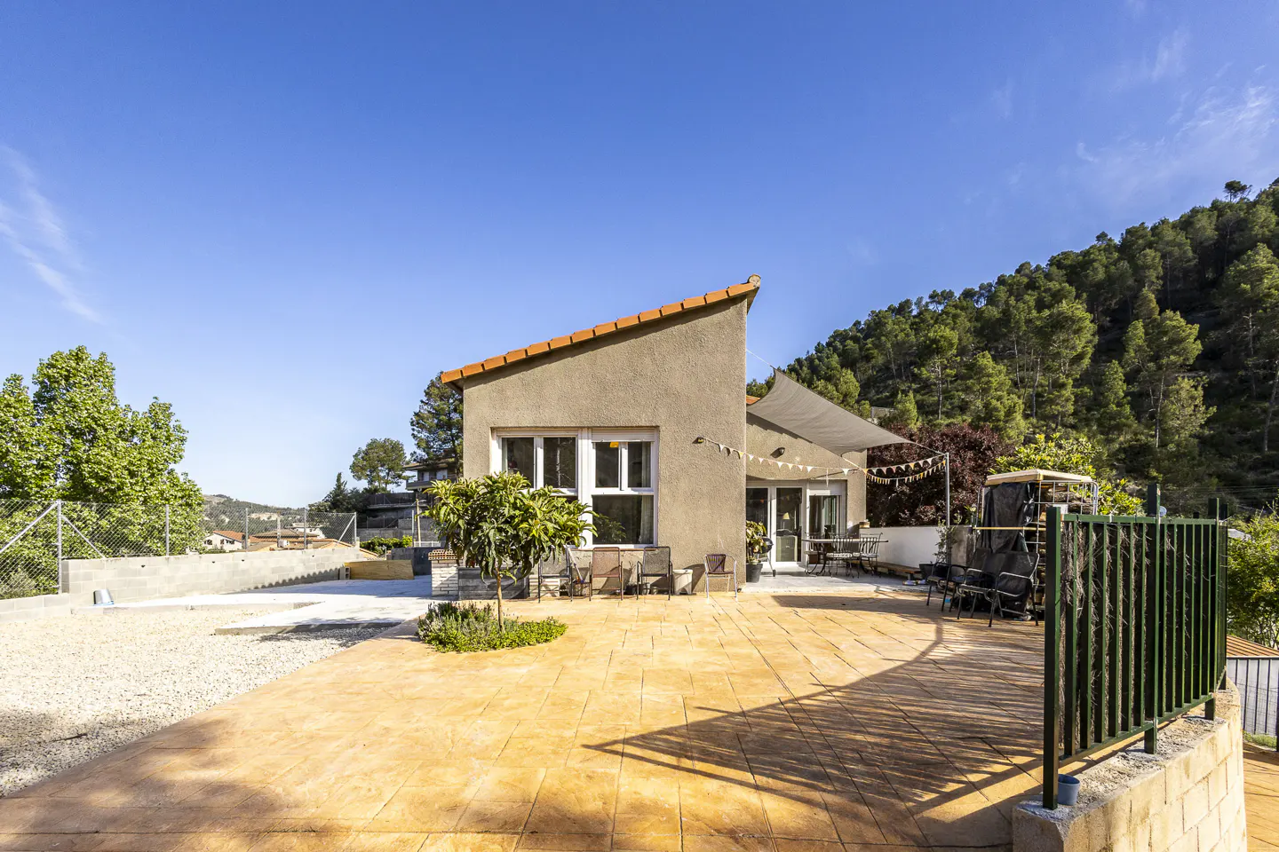 Exterior view of a modern, one-story house with a brown facade, orange roof, and a large patio with outdoor furniture. A green, tree-covered hill rises in the background under a clear blue sky.