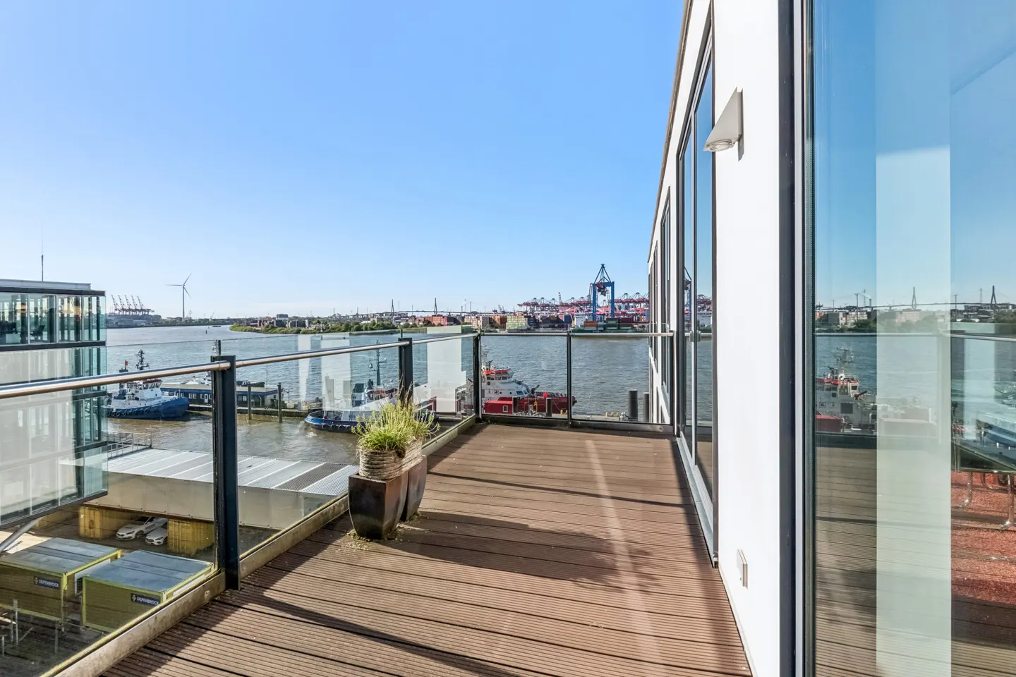Balcony view of a harbor with boats and cranes. The balcony has a wood floor and glass railing. The building is white with large windows.