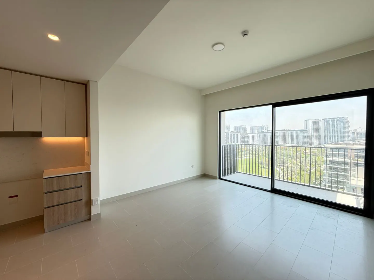 Bright, empty apartment with beige cabinets, drawers, and a large sliding glass door to a balcony with a city view.