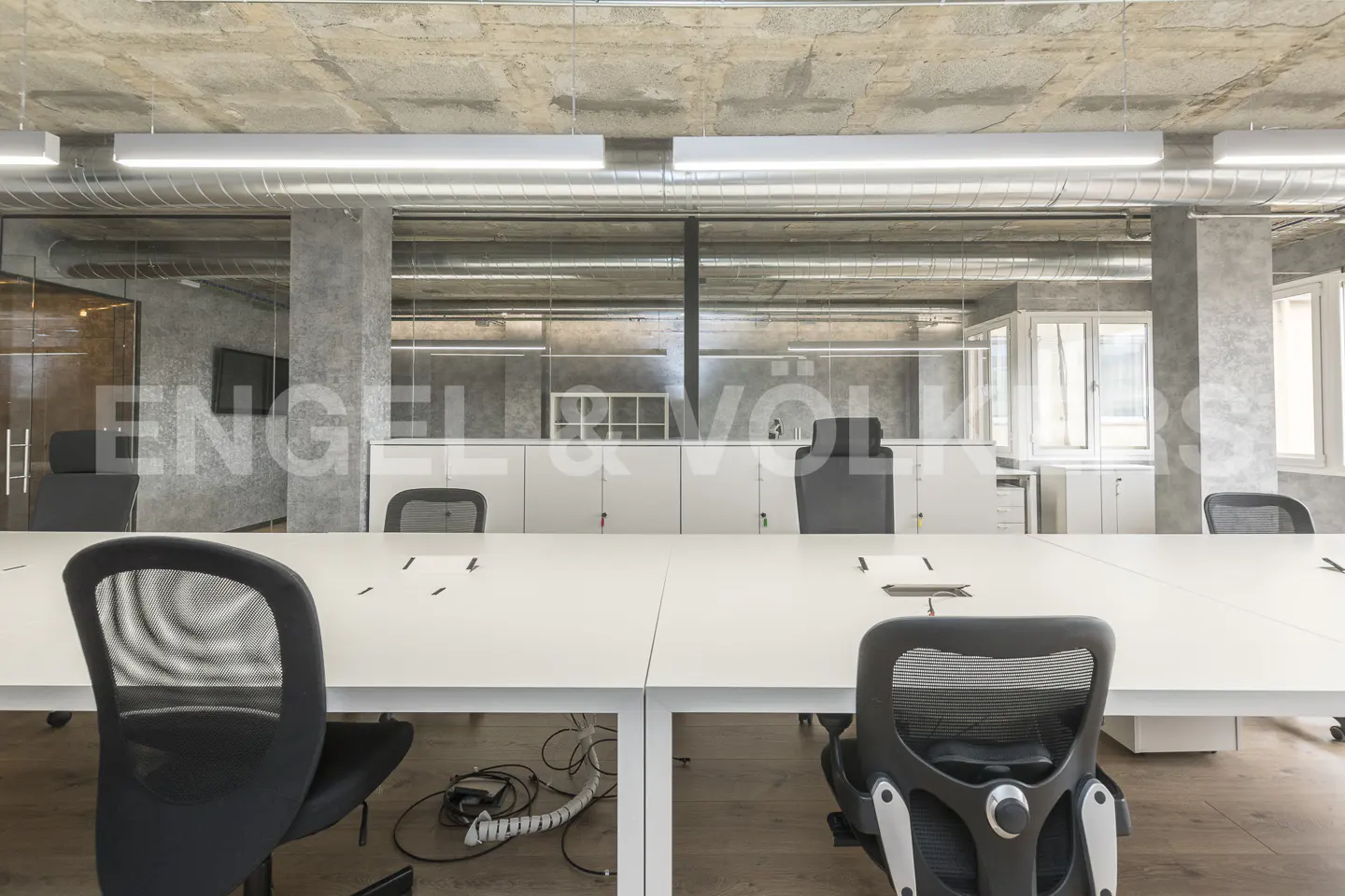 Bright office space with white desks, black chairs, and concrete ceiling. Long fluorescent lights illuminate the room.