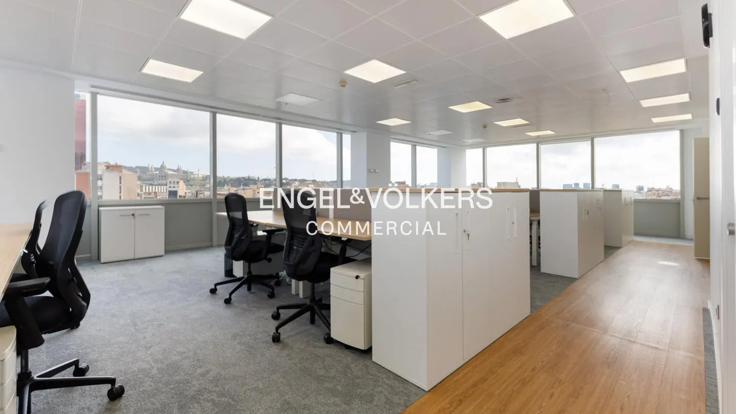 Bright office space with desks, black chairs, and white storage. Large windows offer a city view. Engel & Völkers Commercial logo overlay.