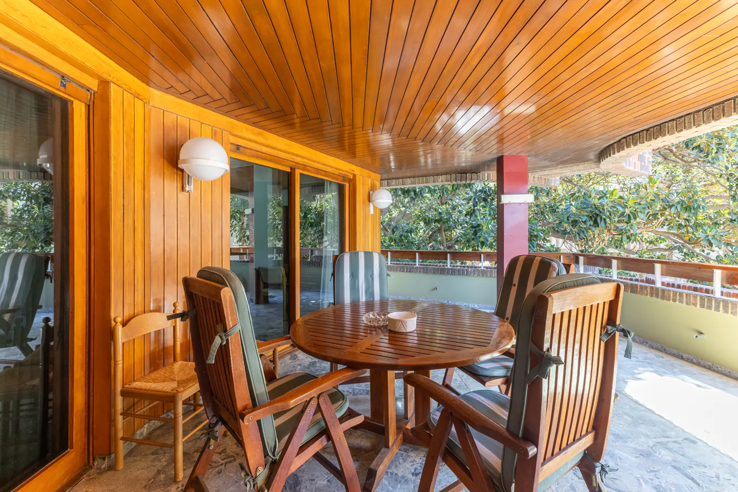 Covered patio with wood ceiling, walls, and furniture. A round table with chairs sits on a stone floor, with trees visible beyond the railing.