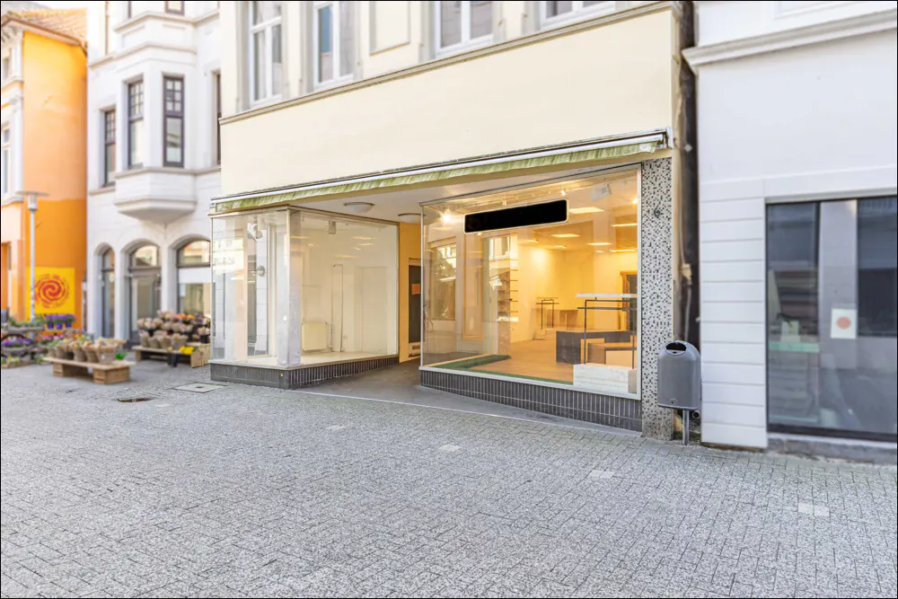 Storefront with large glass windows on a cobblestone street. The interior is brightly lit and mostly empty.