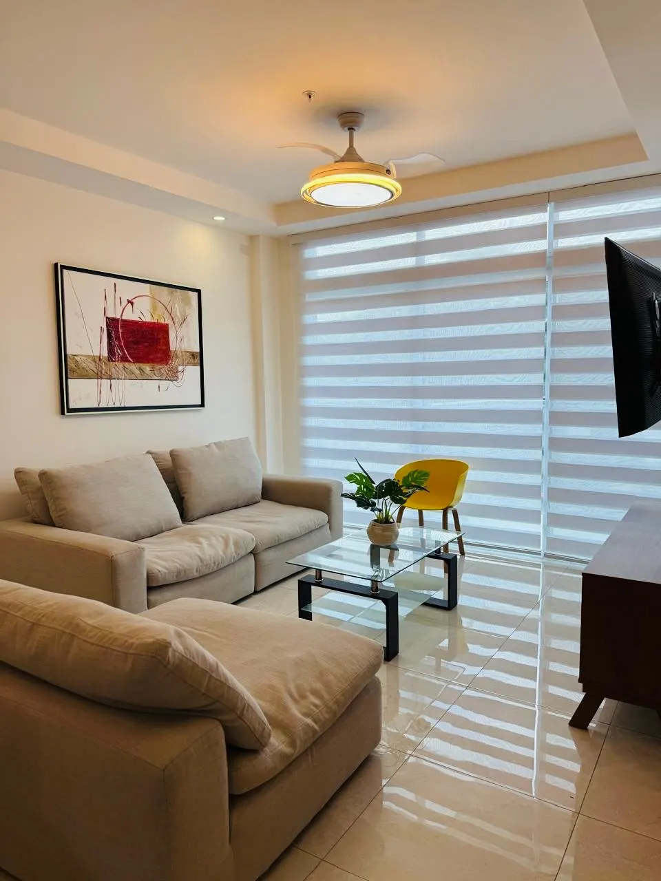 Bright living room with beige sofas, a glass coffee table, and a yellow chair. Abstract art hangs above the sofa. A ceiling fan provides light.