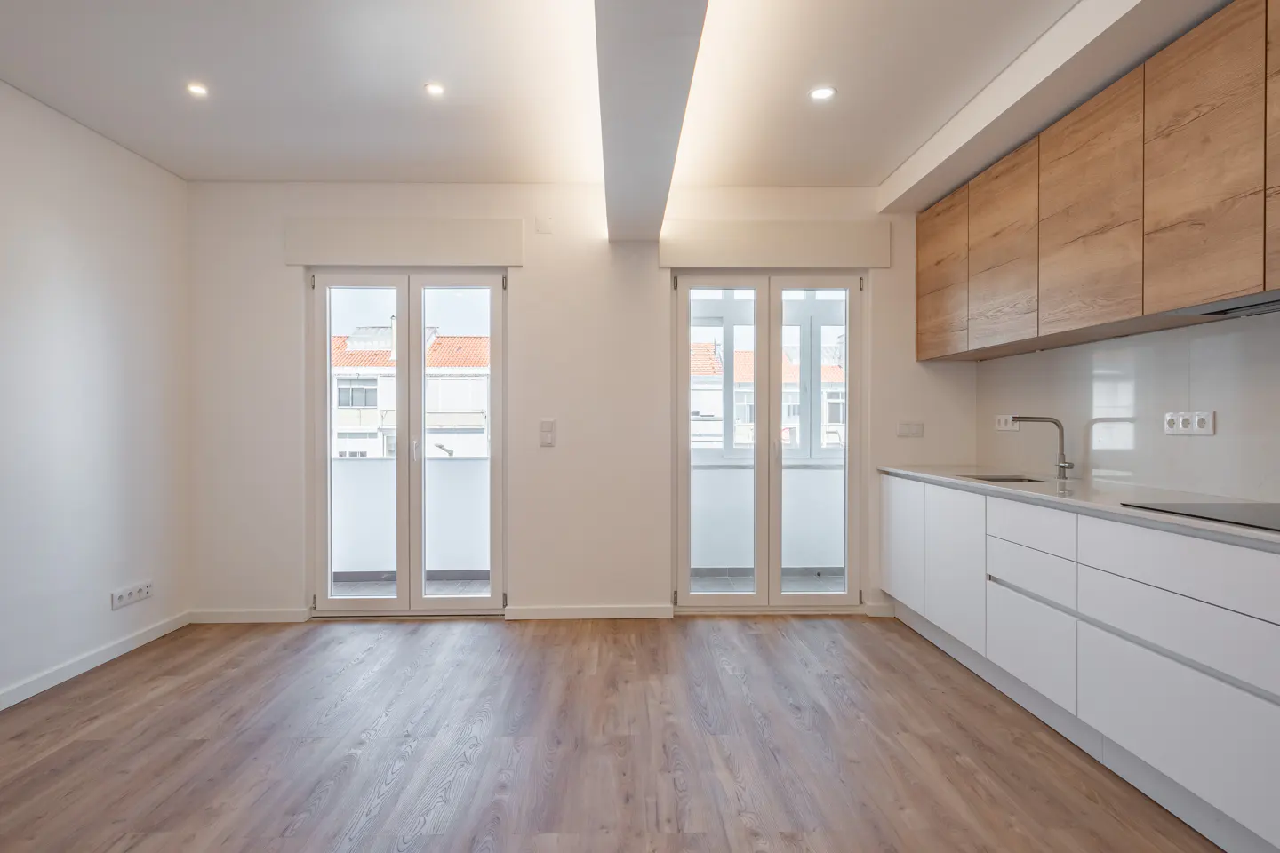 Bright, modern apartment interior with wood floors, white walls, and a kitchen with white cabinets and wood-look upper cabinets. Two sets of white French doors lead to balconies.
