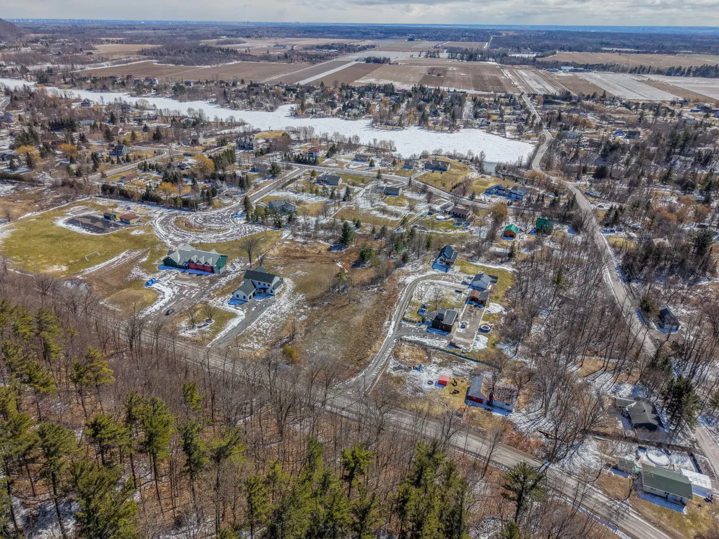 Aerial view of a snow-dusted residential area with houses, trees, and a frozen lake in the background.