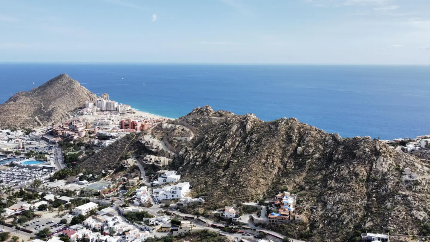 Aerial view of Cabo San Lucas, Mexico, showing mountains, buildings, and the blue ocean under a clear sky.