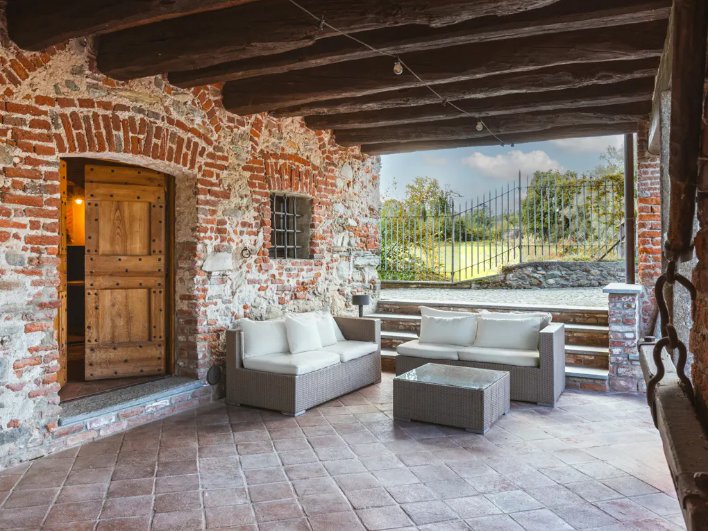 Outdoor patio with brick walls, wooden beams, and tile floor. Two sofas and a glass table sit in front of a gated garden.