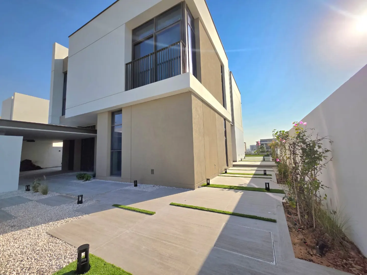 Modern two-story house with white and beige exterior, black balcony, and a walkway with grass accents.