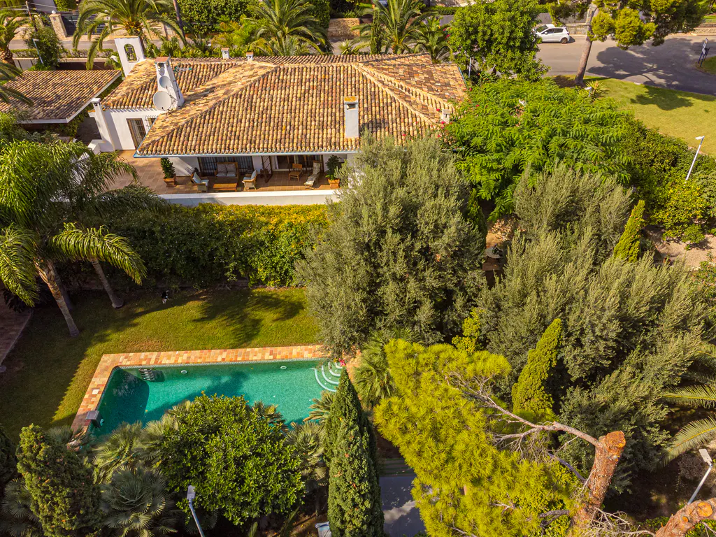 Aerial view of a white villa with a terracotta roof, featuring a swimming pool, lush green garden, and mature trees.