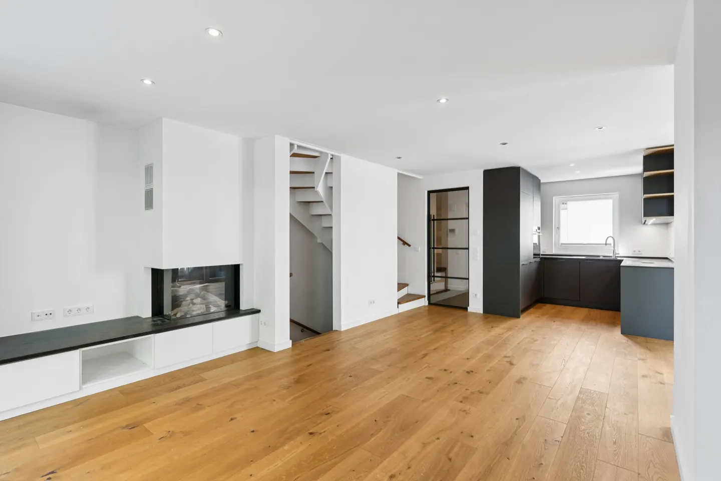 Bright, modern living space with hardwood floors, white walls, a fireplace, stairs, and a black kitchen area.