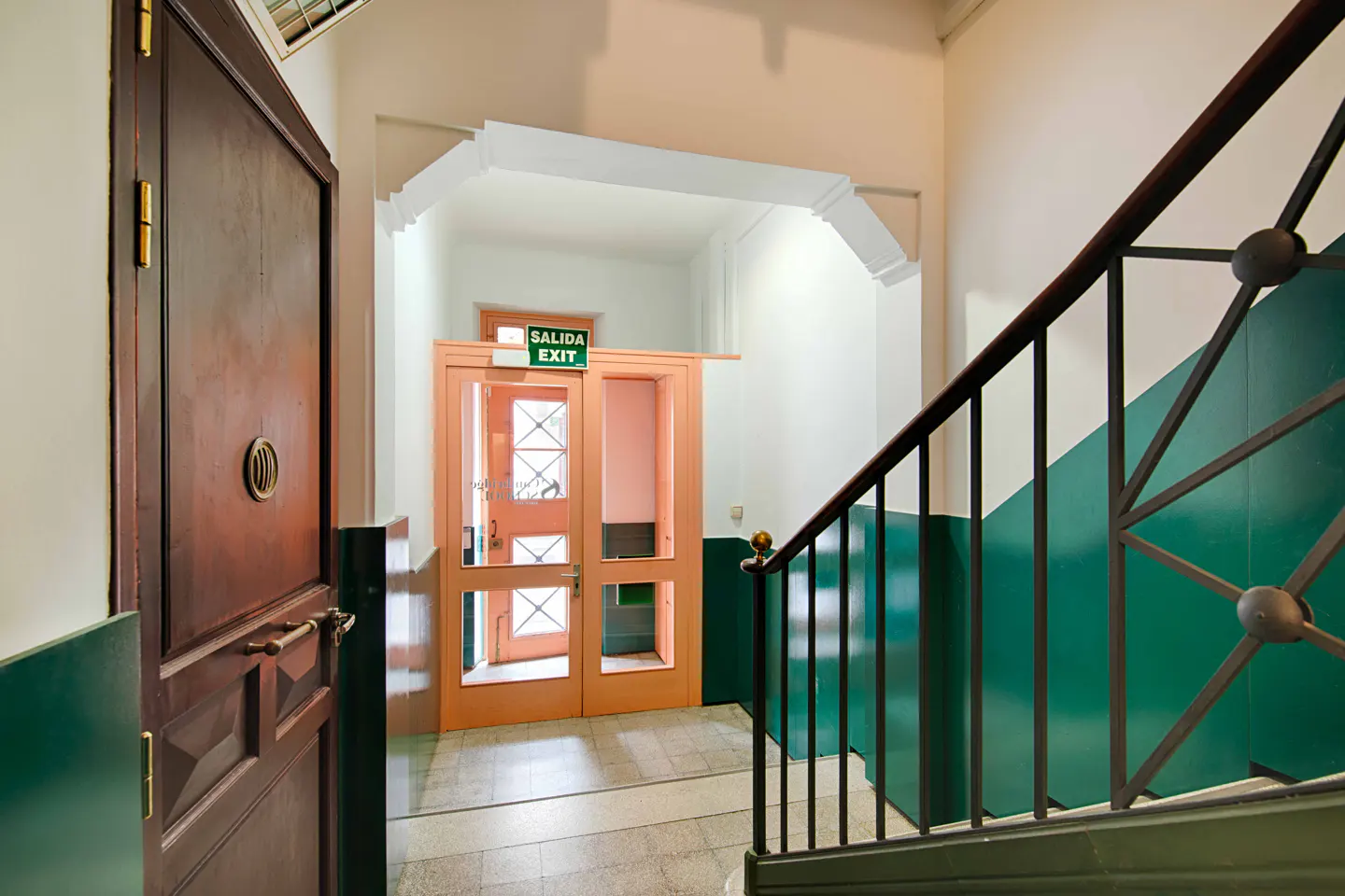 Interior view of a building's hallway with a brown door on the left and an exit door ahead, plus a staircase with green walls.