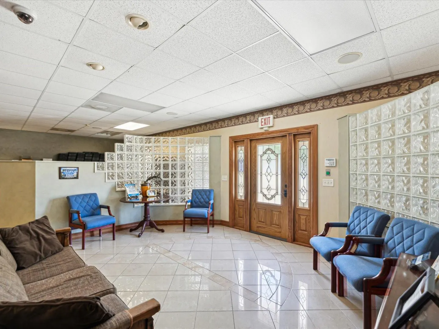 Waiting room with blue chairs, brown sofa, and glass block wall. Wood door with "Exit" sign. Tiled floor and drop ceiling.