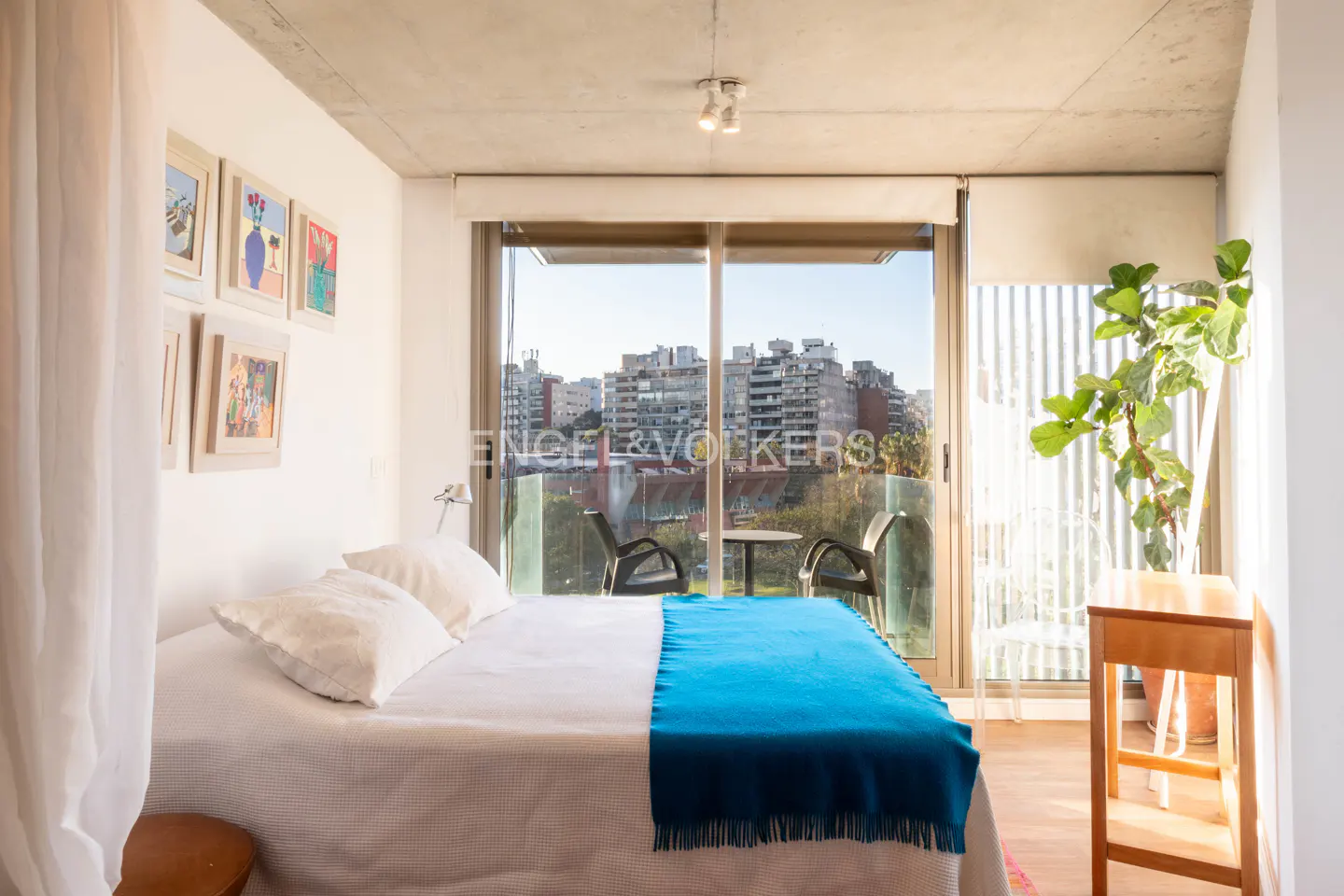 Bedroom with white linens, blue throw, and balcony overlooking city buildings. Artwork on the wall and a potted plant add color.
