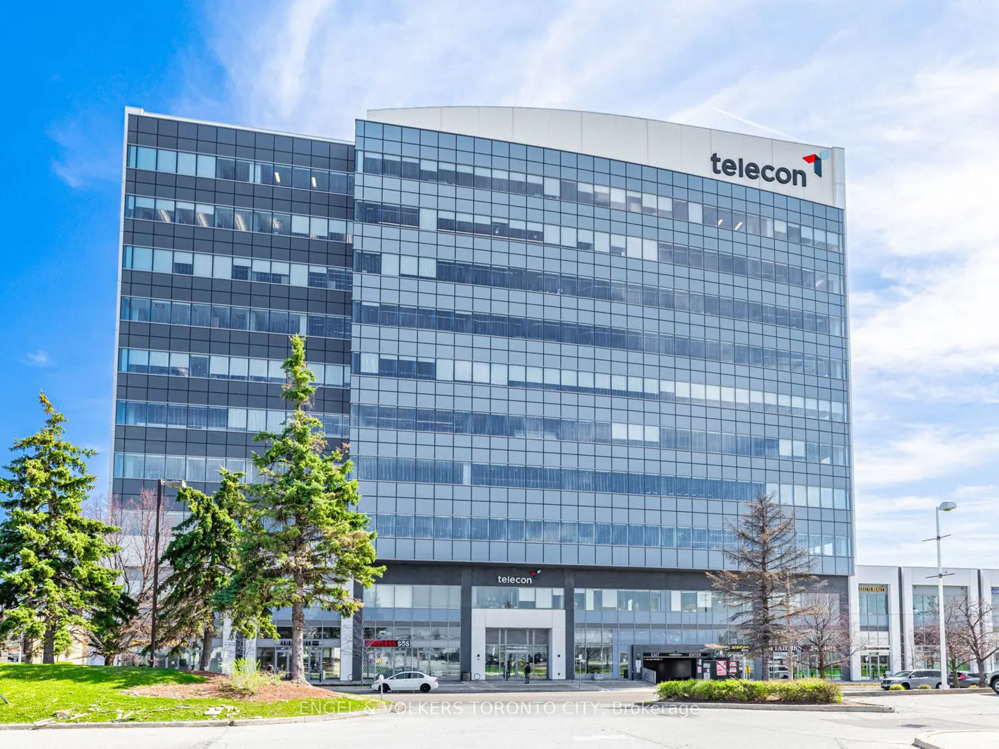 Exterior view of the Telecon building, a modern glass and steel structure under a bright blue sky. Trees line the front of the building.