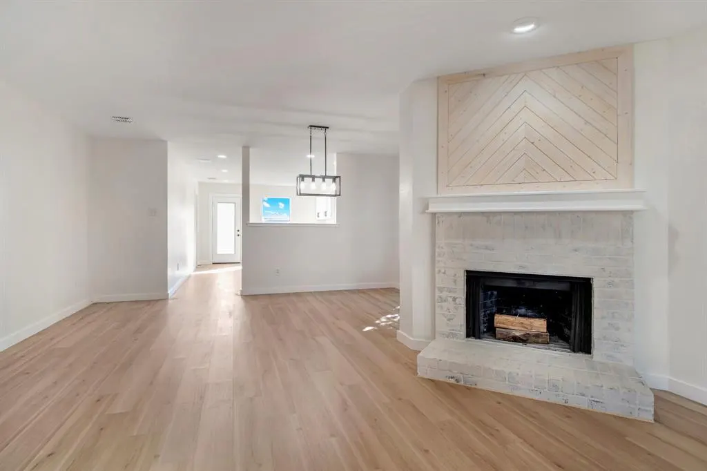 Bright, open-concept living room with light wood floors, white walls, and a brick fireplace with a herringbone wood accent above.