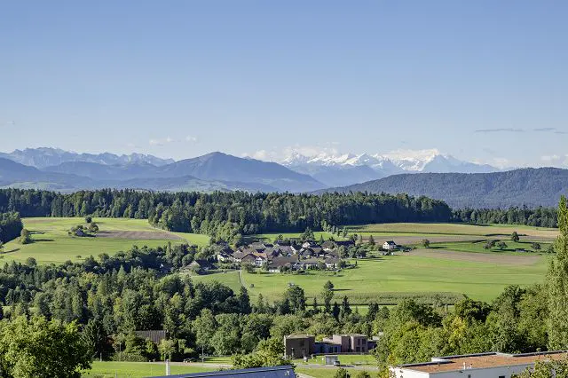 Scenic view of a village nestled in green fields, surrounded by forests and mountains under a clear blue sky.
