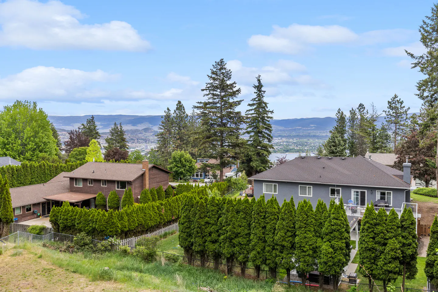 Real estate image showing two houses with green hedges, trees, and a mountain view under a blue sky.