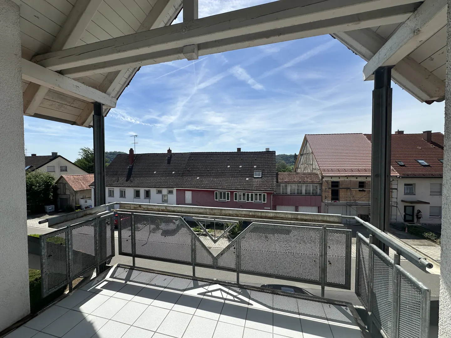 Covered balcony with white tiled floor and metal mesh railing overlooking a street with houses under a bright blue sky.