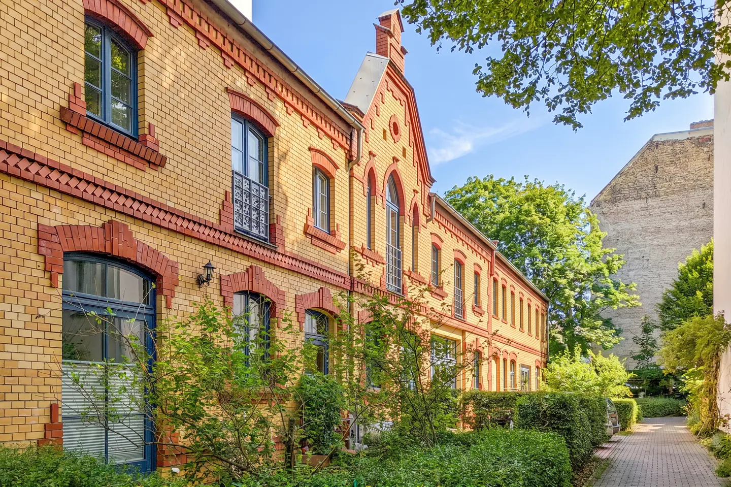 Exterior view of a yellow brick building with red brick trim and blue window frames, surrounded by greenery.