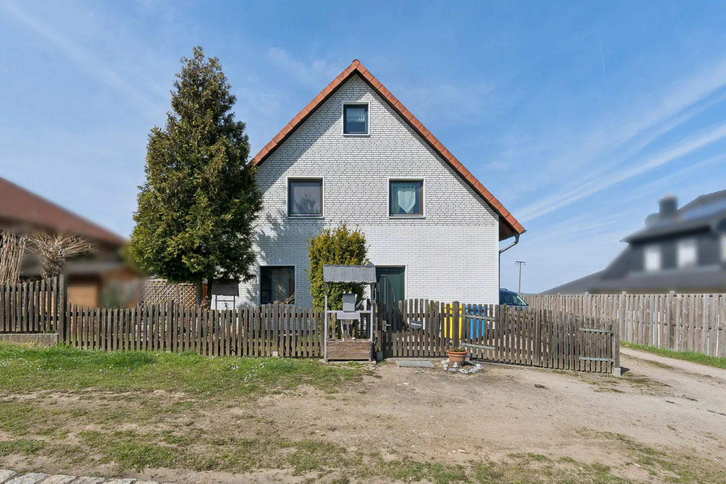 Two-story white brick house with a red roof, surrounded by a wooden fence and green trees under a blue sky.
