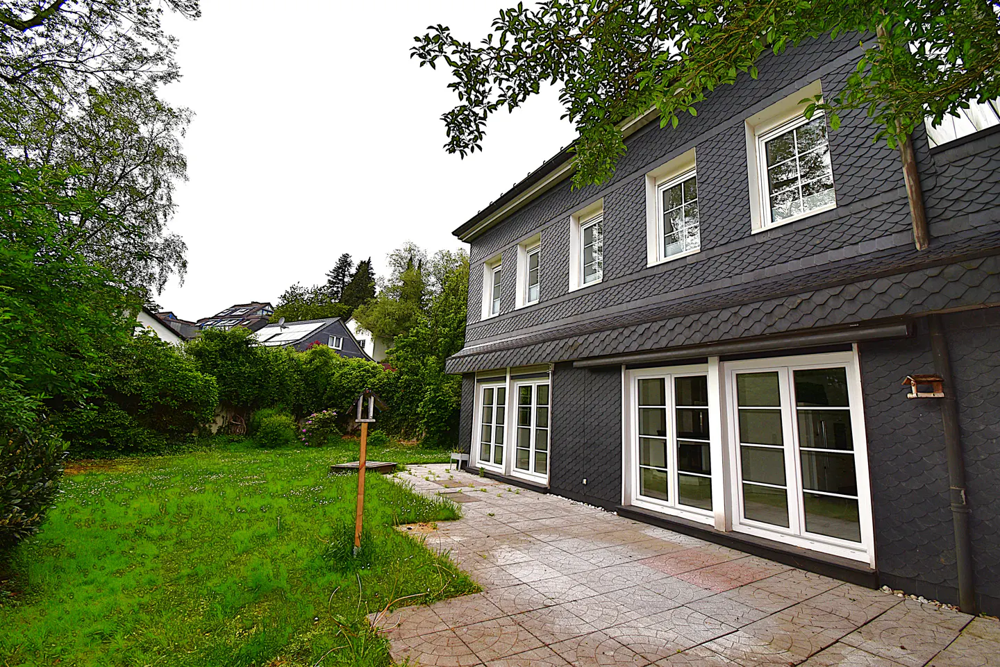 Exterior view of a two-story house with gray shingle siding, white windows, and a stone patio, surrounded by green grass and trees.