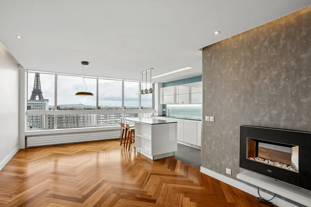 Bright apartment with herringbone wood floors, a white kitchen, and a view of the Eiffel Tower through large windows.