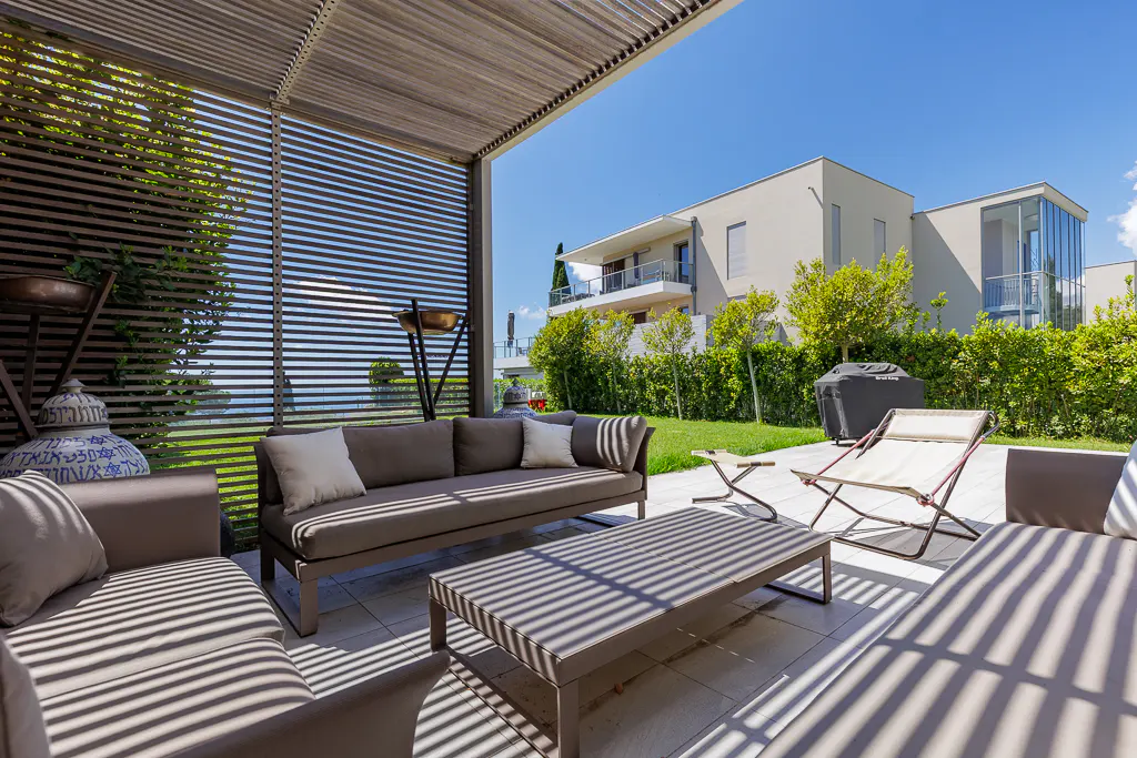 Outdoor patio with gray sofas, a coffee table, and lounge chairs under a pergola. A modern house and green lawn are in the background.