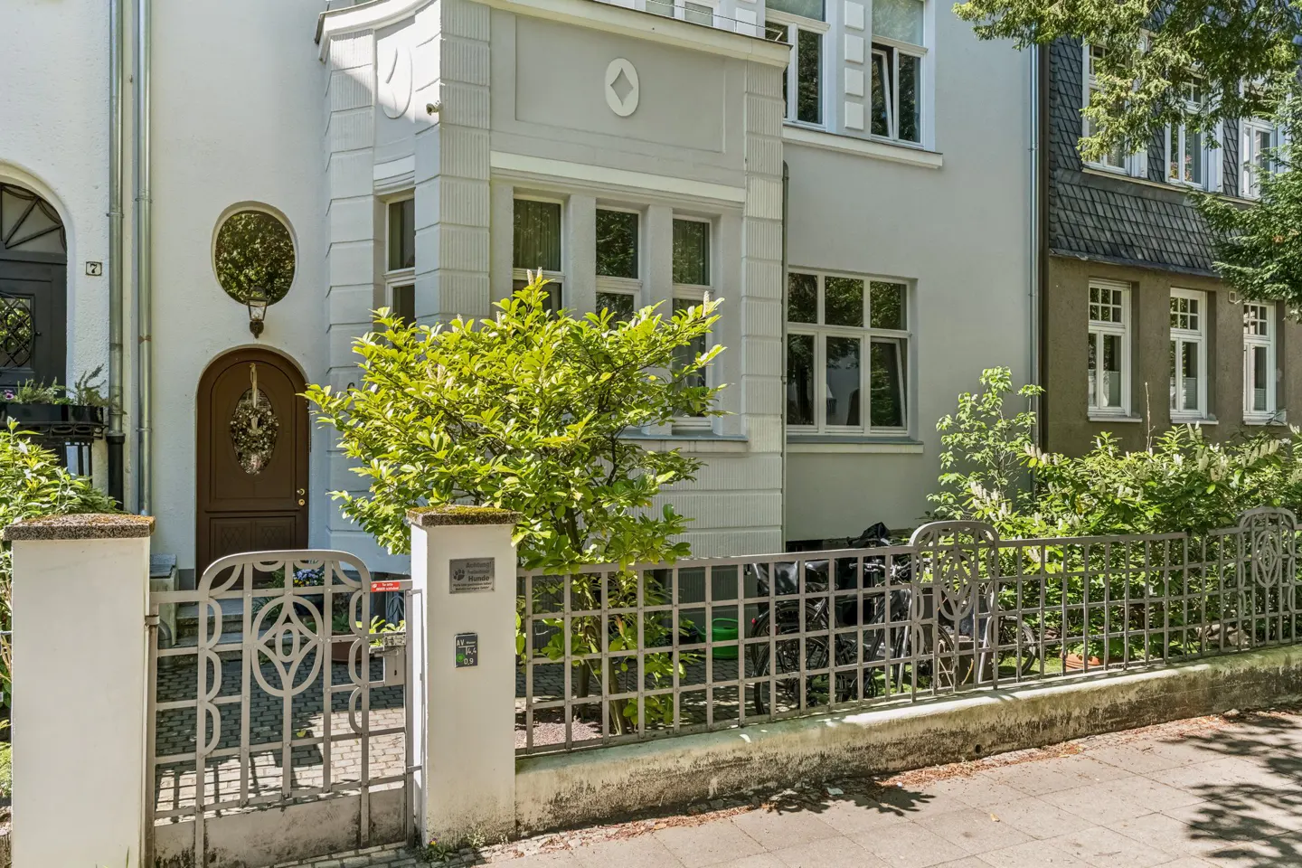 Exterior of a light gray building with a brown arched door and decorative metal gate. Green foliage is visible.